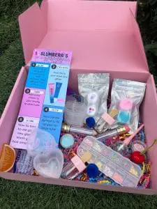 Open DIY Lipgloss Party Box containing various craft supplies, including bottles of glitter, brushes, a syringe, and colorful decorations scattered on grass.