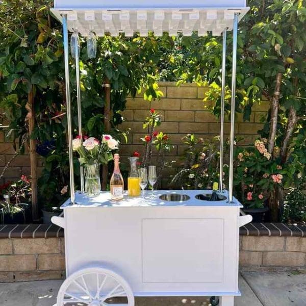 Sentence with Product Name: A white beverage cart adorned with a bouquet of flowers, a bottle of champagne, two glasses, an XL Outdoor Games Addon, and surrounded by lush greenery.