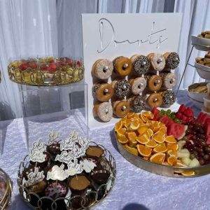 A dessert table adorned with a donut wall, cupcakes, assorted fruits, and various desserts displayed in gold-rimmed trays is perfect for any event. Complement this setup with our XL Outdoor Games Addon to create an unforgettable experience. Our party service ensures everything looks impeccable, making it an ideal choice for kids' party rentals or any celebration requiring top-notch party rental options.