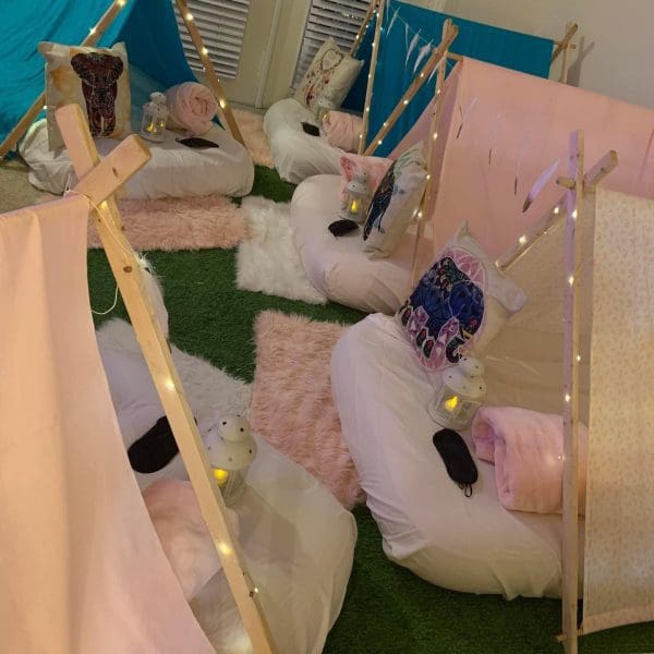 A room with five small indoor teepees, each featuring pillows, blankets, sleeping masks, and small lanterns, all set on a grass-like carpet with a cozy ambiance—perfect for a kids party rental.