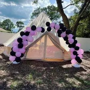 A large 293377074_1435127180244802_8023254540460888526_n tent set up outdoors under a sunny sky, adorned with an arch of black and pink balloons at the entrance.