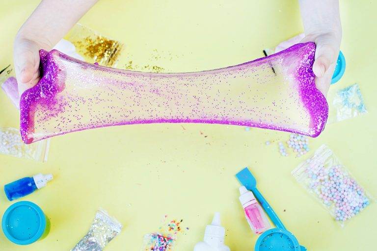 Slime being stretched out with other slime birthday party materials such as glitter, scents, colors and foam balls on the table.