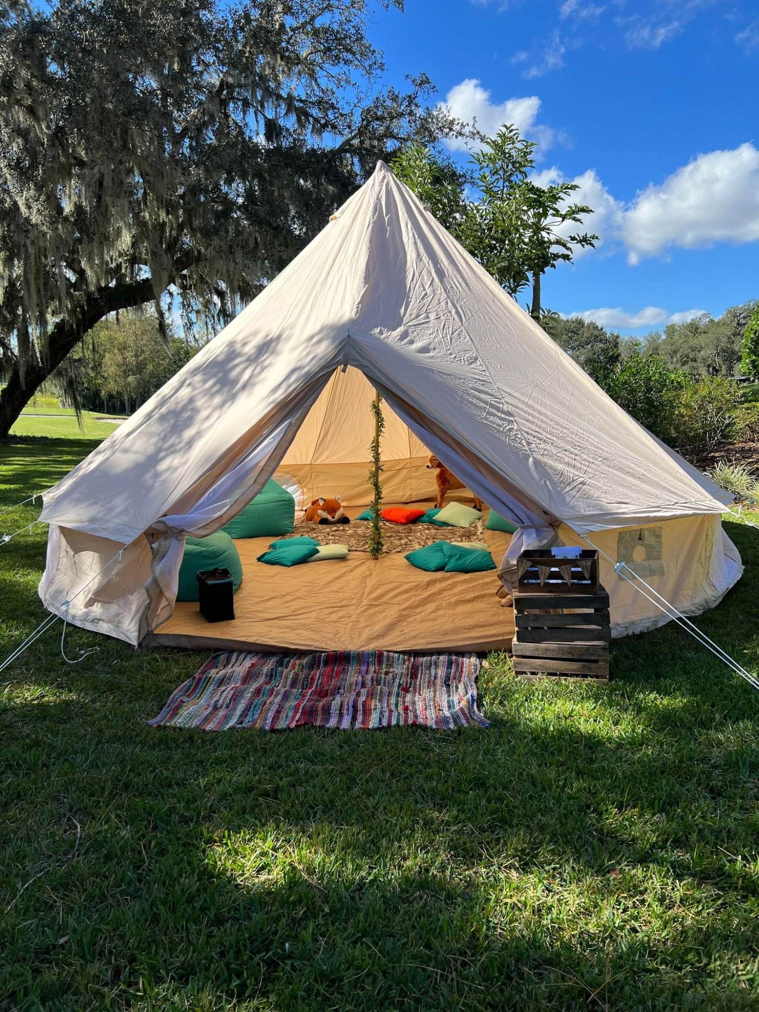 A white tent set up on a grassy field with an open entrance revealing colorful pillows and blankets inside. Perfect for a kids party rental, a small wooden crate and striped rug sit invitingly in front of the entrance.