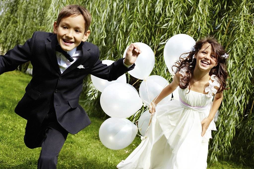 A boy and girl in a wedding dress running with white balloons through a beautiful Lakeland countryside.