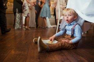 A young child wearing suspenders sits on the floor eating from a bowl, peeking out from under a tablecloth at a kids party rental. Nearby, adults are engaged in conversation while standing.