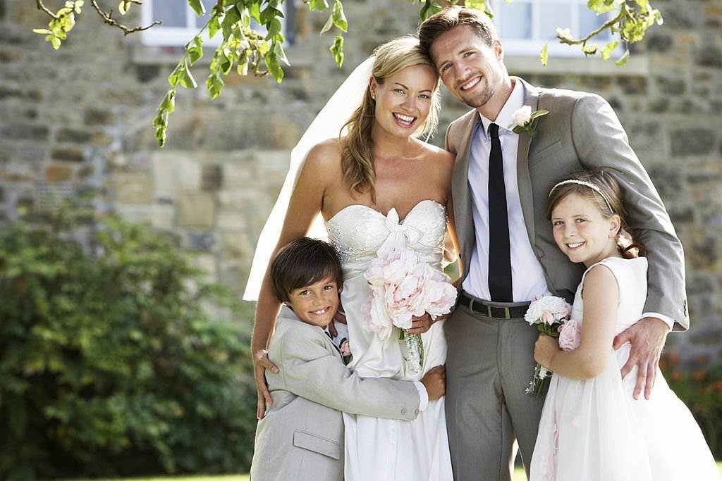 Bride and groom smiling while posing with a young boy and girl outdoors, in front of a stone building. The woman is wearing a strapless white dress, and the man is in a gray suit. A charming teepee stands nearby, adding a whimsical touch to the scene.