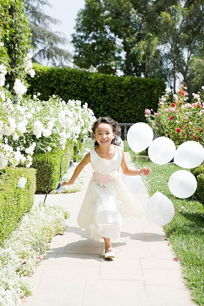 A girl in a white dress running through a garden with balloons, enjoying the glamping experience in a bell tent.