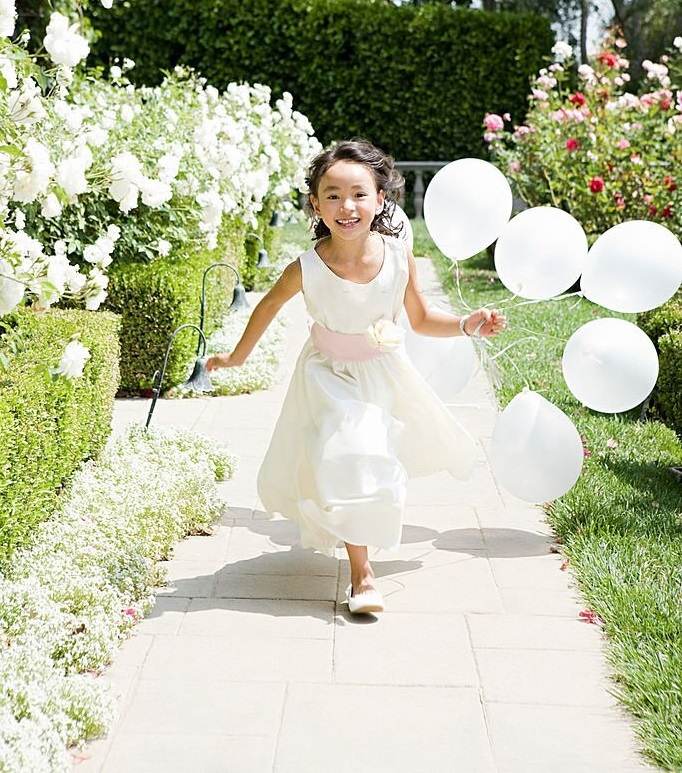 A young girl in a white dress holds white balloons while running on a garden path lined with white flowers and green bushes, her joy reminiscent of the fun at a slime party organized by an exceptional party service.