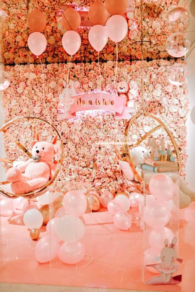 A festive setup for a second birthday party featuring a backdrop of pink flowers, balloons in shades of pink, two large golden rings, build-a-bear teddy bears, and a sign reading A pink and white party with balloons, teddy bears, and a teepee.