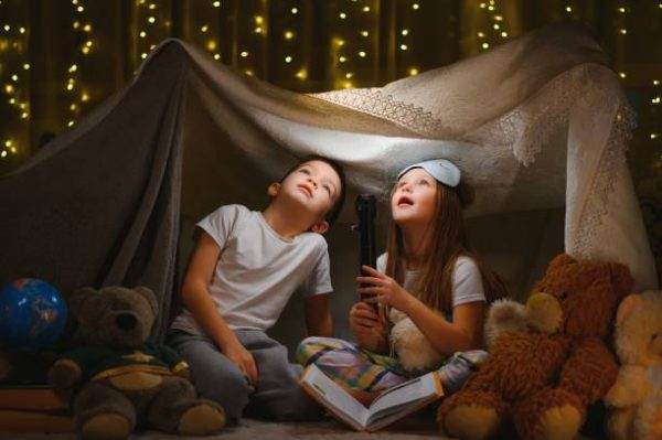 Two children sitting in a teepee with teddy bears and a flashlight.