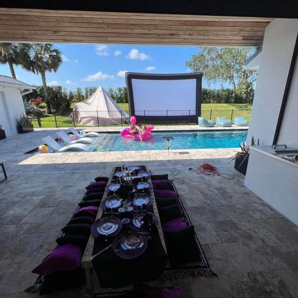 Outdoor dining setup with a long table set for a meal, overlooking a pool with a large screen and inflatable flamingo, in a luxurious backyard perfect for a Lakeland FL party.