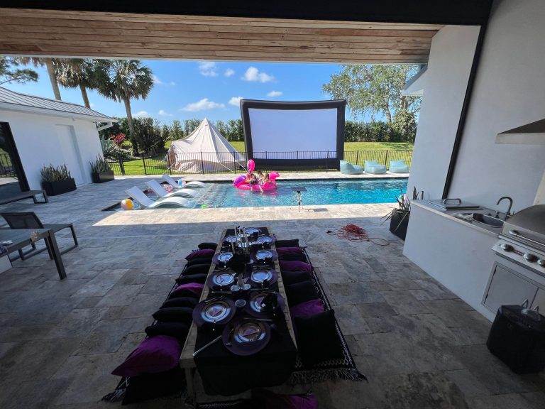 Giant inflatable projector screen sitting beside a luxurious inground pool and bell tent rental. In front of the camera is an outdoor picnic set for a Wednesday Birthday Theme
