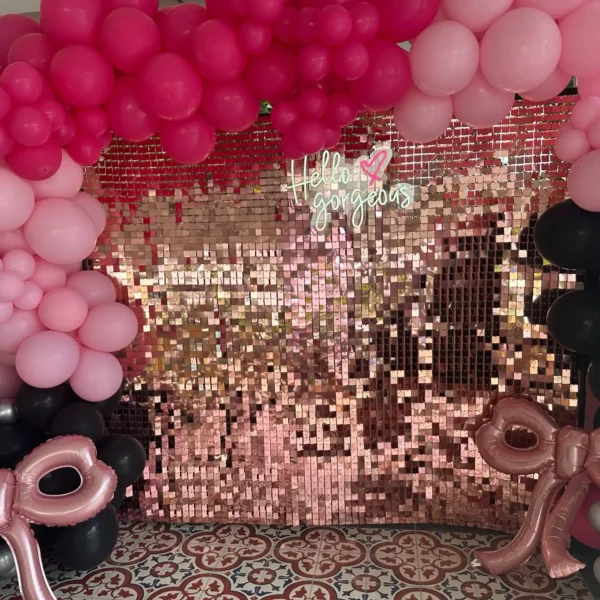 The Shimmer Backdrop Wall Rental, featuring pink and black balloons, metallic bows, a sequin wall, and a "Hello gorgeous" neon sign, creates the ideal photo backdrop for any fashionable celebration.