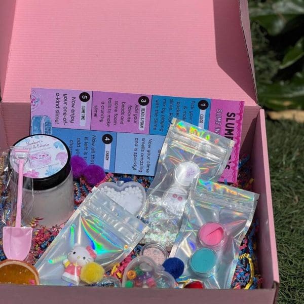 An open box filled with various craft supplies including beads, glitter, and small containers for a Slime Party Box, set on a grassy background.