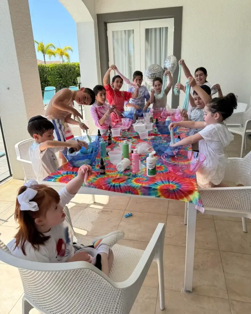 A group of children make slime with various supplies on a tie-dye table outdoors, next to a Shimmer Backdrop Wall Rental that adds sparkle and excitement to their creative activity.