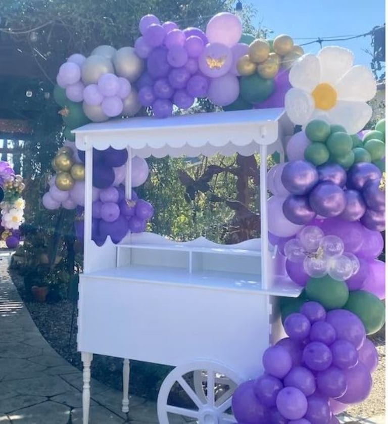 An Elegant White Snack or Candy Cart Rental adorned with vibrant purple and green balloons, ideal for a celebration in Lakeland.