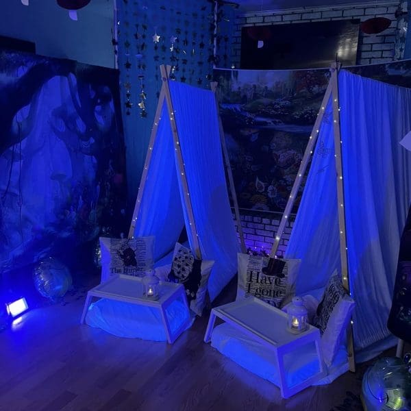 Indoor camping setup with two teepees, fairy lights, and Alice in Wonderland-themed decorations under a blue light.