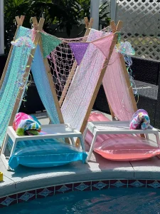 Enchanting poolside retreat showcasing stylish Teepee Cabanas with elegant wooden frames, beautifully draped in soft pastel hues of blue, pink, and purple fabric. Each chic cabana features a delicate net canopy and a lively pennant banner fluttering in the breeze. Beneath these inviting shelters are plush inflatable lounge pads in complementary shades of blue and pink, each meticulously topped with neatly rolled towels for ultimate relaxation. The serene ambiance is further enhanced by the luxurious pool’s intricately designed decorative tiles. Perfect for a tranquil escape or vibrant summer gathering!