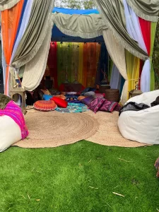 Colorful outdoor tent with draped fabrics and an array of cushions on a circular mat, designed for a **Dream Catcher Themed Party**, set against a backdrop of green grass.