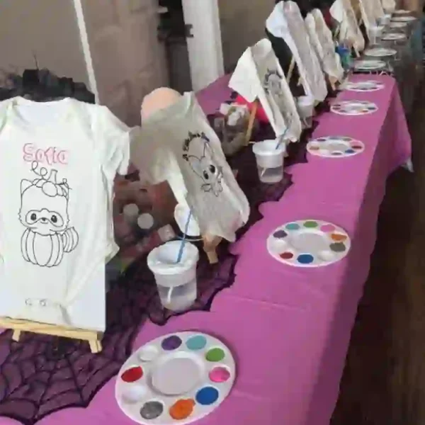 Join the Ultimate Baby Shower Celebration with a Crafty Onesie Painting Party! Experience a vibrant and captivating scene as adorable baby onesies, featuring charming cartoon designs of animals and beloved characters, are artfully displayed on easels. These creative setups rest on a stylish pink tablecloth, complete with artist palettes and brushes immersed in water cups, inviting you to unleash your artistic flair. Let your imagination run wild as you paint and personalize these tiny outfits in a fun-filled gathering. Behind this colorful arrangement lies an elegantly-lit hallway that adds depth to this delightful artistic experience. Perfect for expecting parents looking to add unique memories to their baby shower festivities!
