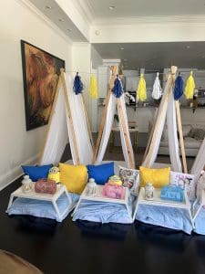Three teepee-style tents are set up indoors, adorned with blue and yellow pillows, trays with lanterns, and plush toys in front. Decorative tassels hang from the tents, while an abstract painting graces the wall. The setup feels like a whimsical auto-draft of a cozy dream. 1 Three teepee-style tents are set up indoors, adorned with blue and yellow pillows, trays with lanterns, and plush toys in front. Decorative tassels hang from the tents, while an abstract painting graces the wall. The setup feels like a whimsical auto-draft of a cozy dream.
