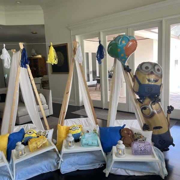 Three indoor tents with pillows and blankets, each with a tray holding a lantern, teddy bear, and gift bag. An AUTO-DRAFT balloon stands near the rightmost tent, next to the Minion-themed balloon.