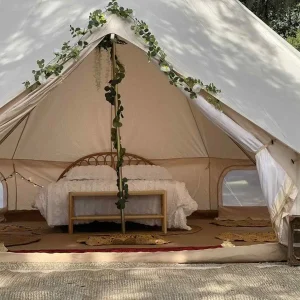 A glamping tent with an open entrance reveals a bed adorned with white bedding, making it feel like a cozy home. The tent is decorated with green foliage and fairy lights, creating an enchanting atmosphere.