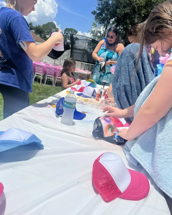 Children and adults are gathered around an outdoor table with paint supplies, decorating caps. A woman in a blue dress assists them. The sunny scene is adorned with trees and luxury outdoor furniture, creating a perfect setting for their Luxury Soft Play Packages.