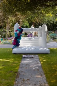 An Event Photography Package features an inflatable white bouncy castle adorned with a delightful cluster of purple, blue, and pink balloons, set on a grassy area with a stone pathway leading up to it. The enchanting backdrop of trees and garden adds the perfect touch for capturing memorable moments. 1 An Event Photography Package features an inflatable white bouncy castle adorned with a delightful cluster of purple, blue, and pink balloons, set on a grassy area with a stone pathway leading up to it. The enchanting backdrop of trees and garden adds the perfect touch for capturing memorable moments.