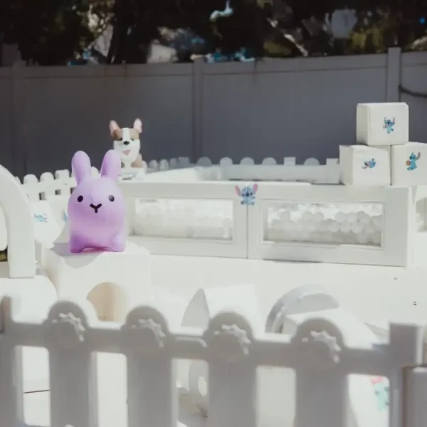 A delightful scene is created with a purple bunny toy placed inside a small white fenced enclosure, accompanied by white structures and a smaller toy in the background, making it ideal for showcasing Event Photography Packages.