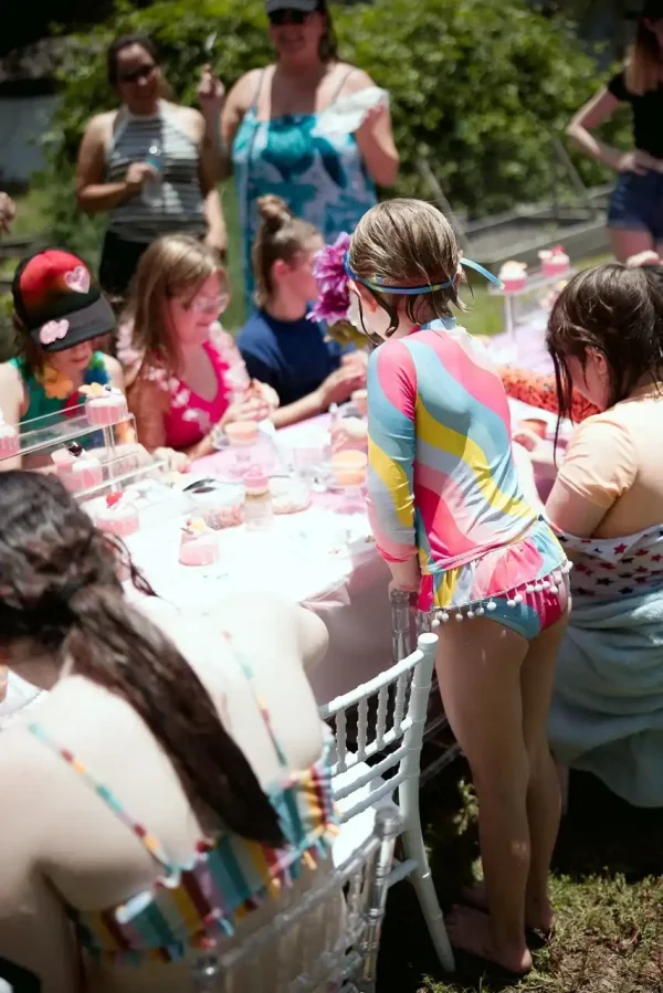 A group of children in swimsuits are gathered around a decorated table outdoors, engaging in activities, while several adults stand observing in the background, capturing moments that could be part of the Event Photography Packages.