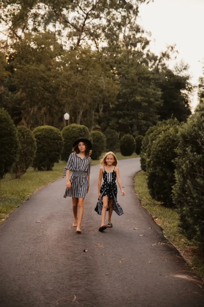 Two young girls revel in a magical day organized by Slumberr, the premier luxury children's party planner. As they walk barefoot along a sun-dappled paved path, their laughter blends with the serene ambiance of the garden. The taller girl exudes elegance in her chic striped dress and oversized wide-brimmed sun hat, while her friend dazzles in a vibrant patterned sleeveless dress. Surrounded by lush greenery and immaculately trimmed bushes, they embody youthful joy and carefree adventure amidst nature's beauty.
