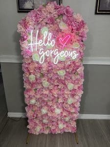 Immerse yourself in the enchanting beauty of a stunning floral wall display, featuring an opulent arrangement of lush pink and white roses. Perfect for adding a touch of elegance to any setting, this vibrant floral backdrop is accentuated by an eye-catching neon sign that reads "Hello Gorgeous," accompanied by a charming pink heart symbol. The abundance of flowers are meticulously arranged on a chic rectangular board, set against a sophisticated beige wall adorned with stylish gray wainscot paneling. At the base, the polished wooden floor adds warmth and contrast to this captivating scene. Ideal for photo ops or memorable events, this dazzling rose display effortlessly combines luxury and charm.