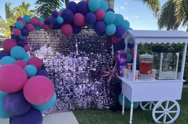 Immerse yourself in a vibrant celebration scene featuring a stunning balloon arch bursting with hues of pink, purple, and teal. The festive ambiance comes alive with a mesmerizing sparkling backdrop that adds an enchanting touch. To the right, you'll find an elegant white serving cart from Slumberr showcasing a stylish drink dispenser complemented by clear cups, ready for guests to enjoy refreshing beverages. Set against this captivating display are lush palm trees and verdant grass under the expansive clear blue sky, perfectly embodying the essence of an outdoor party paradise.