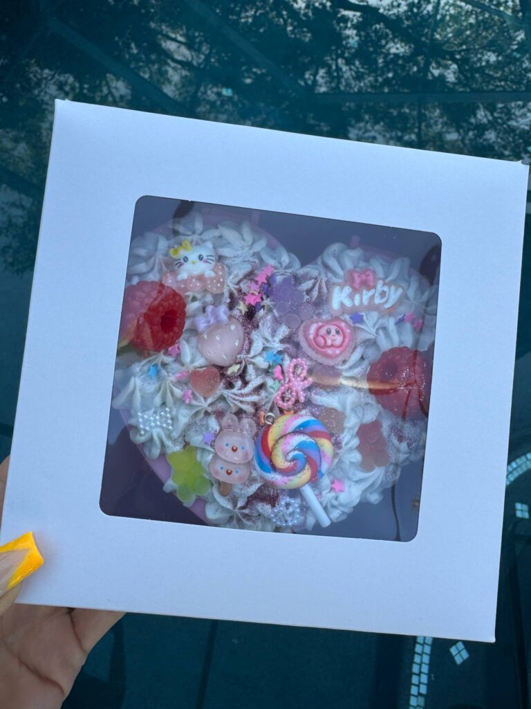 A heart-shaped cake with colorful decorations, including candies, pearls, and a pink "Kirby" topper, sits in a white box with a clear window—perfect inspiration for your next DIY Jewelry Box or Activity Station creation.