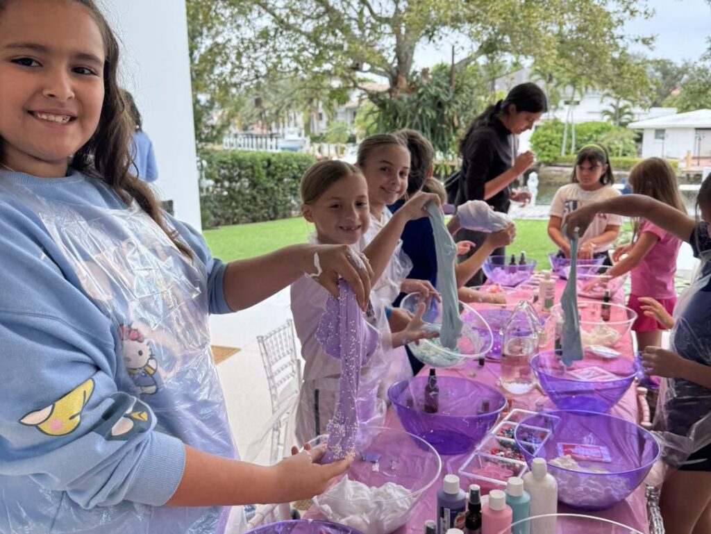 A group of children gather around a table at a party shop, making slime with purple bowls, craft materials, and bottles, while smiling and showing their creations.