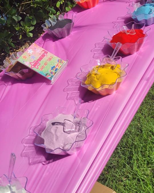 Several bowls filled with different colors of powder, each with a scoop, are arranged on a pink tablecloth outdoors next to a colorful kids activity box.