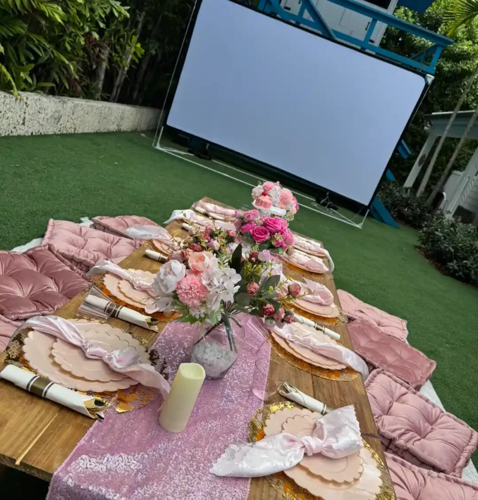 A picnic-style table set with pink cushions, floral centerpieces, and a sequin runner, arranged outdoors on grass in front of a large blank projector screen—perfect for gatherings in Central Florida or styled by your favorite party shop.