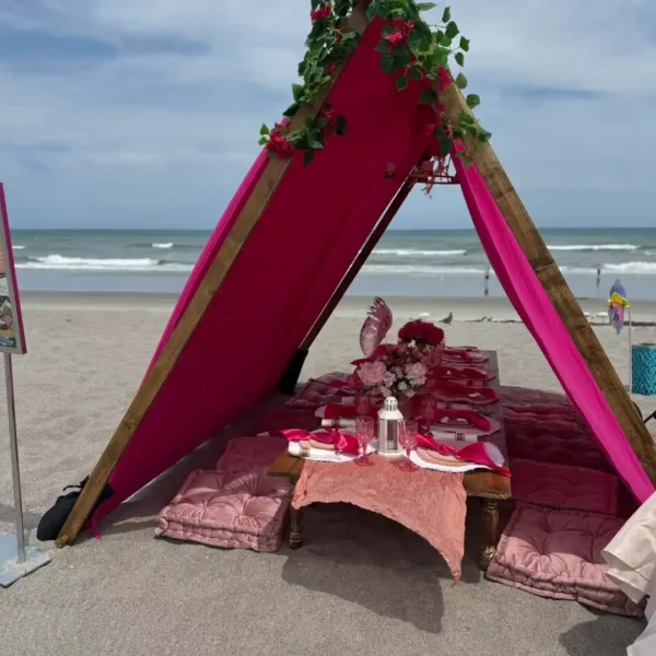 An Extra Large Teepee Rental is set up on a sandy beach with a low table, pink cushions, tableware, and floral decorations under the tent. The ocean can be seen in the background.