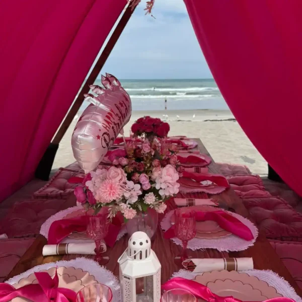 An Extra Large Teepee Rental is set up for a beachside picnic, featuring a decorated table with pink flowers, plates, napkins, a birthday balloon, and a white lantern, all with the ocean as the backdrop.