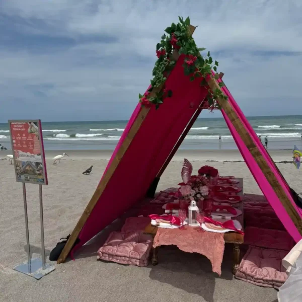 An Extra Large Teepee Rental with floral decorations covers a low dining table on a sandy beach, surrounded by seating cushions and an ocean backdrop.