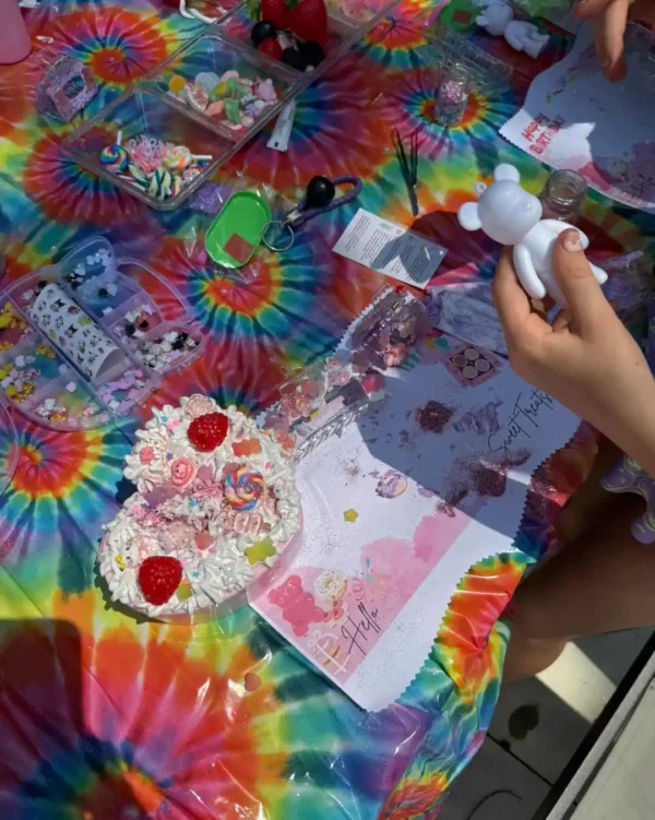 At a tie-dye covered table with craft supplies and charms, someone decorates a white bear figurine beside an Extra Large Teepee Rental—ideal for unique gatherings or special event teepee rentals.