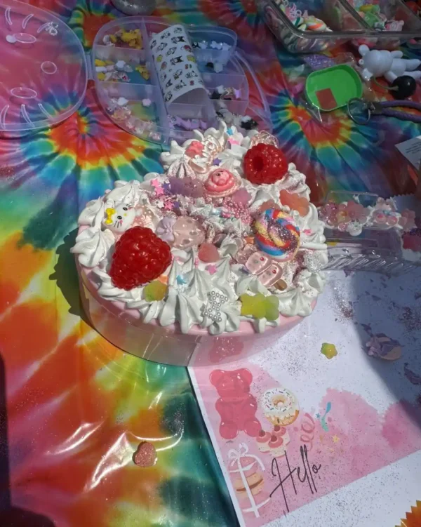 A heart-shaped cake with candies, sprinkles, and cream sits on a tie-dye tablecloth next to craft supplies, pink illustrated paper, and an Extra Large Teepee Rental set up for a cozy celebration.