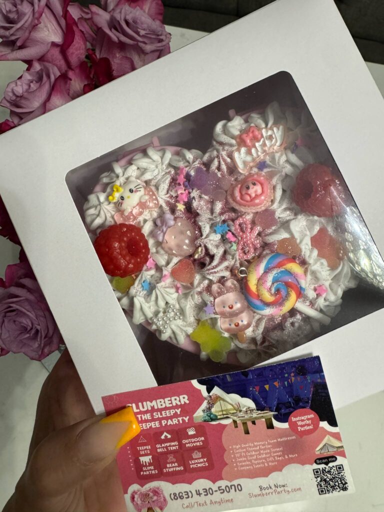 A hand holds a boxed, heart-shaped cake decorated with colorful candies and animal toppers, next to a business card for a party planning service featuring DIY Jewelry Box kits. Pink flowers are in the background.
